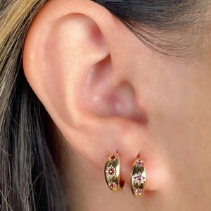 May include: A pair of gold hoop earrings with small pink flower accents. The earrings are worn in the earlobe.