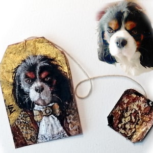 May include: A hand-painted tea bag tag featuring a Cavalier King Charles Spaniel dog wearing a gold and white costume. The tag is made of paper and has a gold background. The dog is looking directly at the viewer with a serious expression.