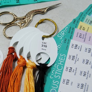 May include: A close-up of embroidery floss color swatches with thread wound around white plastic bobbins. The bobbins are labeled with the brand "CXC" and a color number. The floss colors include baby blue, light green, yellow, pink, white, orange, red, and black. The image also includes a gold pair of scissors, a turquoise measuring tape, and a turquoise and white card with the text "SIRIOUS STITCHES" and a color chart.