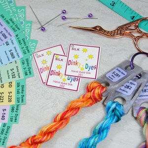 Puede incluir: Primer plano de una colección de hilo de bordar de seda teñido a mano en varios colores, incluyendo naranja, rojo, amarillo, azul y verde. El hilo está dispuesto en espiral y está acompañado de pequeñas etiquetas con el texto "Dinky Dyes" y "Seda teñida a mano".