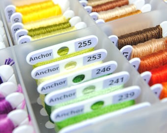 May include: A translucent plastic storage container filled with organized embroidery floss bobbins. Each bobbin is wrapped in colorful thread and features a white label printed with the brand name Anchor and specific numerical color codes.