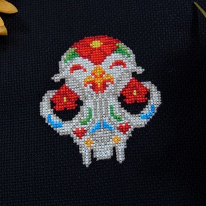 May include: A colorful cross-stitch design of a sugar skull with red, white, blue, green, and yellow embroidery floss. The skull is smiling and has flowers on the top of its head.