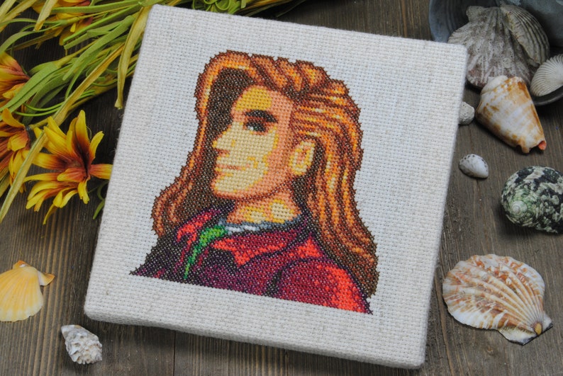 May include: Cross-stitch artwork of a person with long brown hair, wearing a red and green jacket. The artwork is framed on a beige canvas.