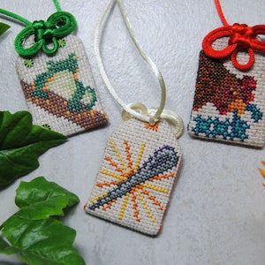 May include: Three embroidered Japanese omamori charms with different designs. One features a dragonfly, another a bird, and the third a stylized sun. Each charm is rectangular, with a loop for hanging, and is made with colorful cross-stitch.
