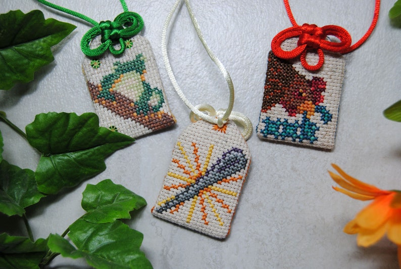 May include: Three embroidered Japanese omamori charms with different designs. One features a dragonfly, another a bird, and the third a stylized sun. Each charm is rectangular, with a loop for hanging, and is made with colorful cross-stitch.