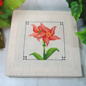 Puede incluir: Un dise&ntilde;o floral de punto de cruz con un lirio rojo y naranja con hojas y tallo verdes, enmarcado en negro sobre tela beige. La labor de aguja es detallada y la pieza es cuadrada.