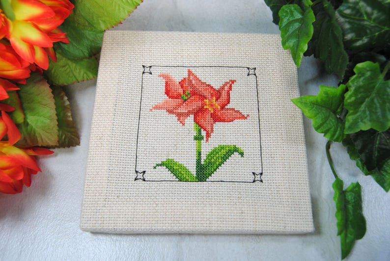 Puede incluir: Un dise&ntilde;o floral de punto de cruz con un lirio rojo y naranja con hojas y tallo verdes, enmarcado en negro sobre tela beige. La labor de aguja es detallada y la pieza es cuadrada.