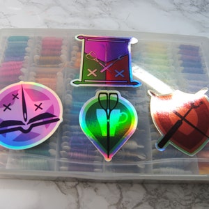 May include: Four iridescent holographic stickers with different designs. The stickers feature a book with a needle and thread, a cup with scissors, a scroll with crossed needles, and a shield with crossed needles.