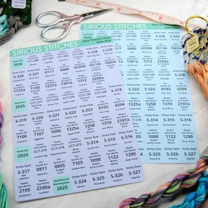 May include: An assortment of colorful embroidery floss and thread, with coordinating color cards labeled "SIRIOUS STITCHES". A pair of scissors and a measuring tape are also present, suggesting a crafting or sewing project.
