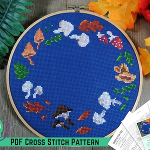 May include: A PDF cross stitch pattern featuring a blue background with a wreath of mushrooms, leaves, and acorns. The pattern is displayed in a wooden embroidery hoop.