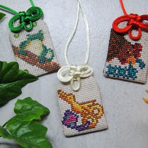 May include: Three embroidered Japanese omamori charms with different designs. One features a green frog, another a brown bear, and the third a golden genie lamp. Each charm has a decorative knot and cord in green, red, and white.