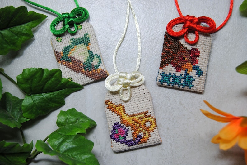 May include: Three embroidered Japanese omamori charms with different designs. One features a green frog, another a brown bear, and the third a golden genie lamp. Each charm has a decorative knot and cord in green, red, and white.
