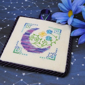 May include: Square cross-stitch art featuring a crescent moon in shades of purple and blue, with floral accents in green, yellow, and blue. The artwork is framed with a dark purple rope trim and a matching ribbon loop for hanging. The background is a dark blue fabric with a constellation pattern.