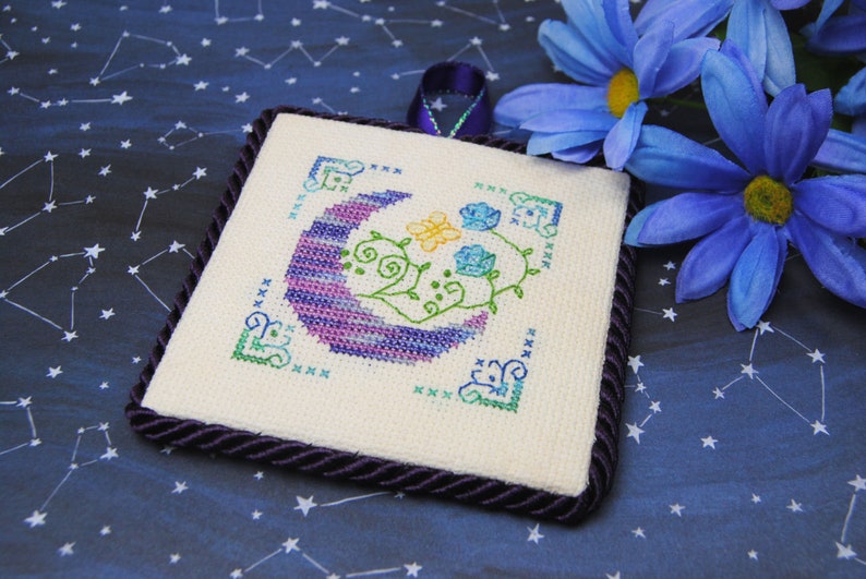 May include: Square cross-stitch art featuring a crescent moon in shades of purple and blue, with floral accents in green, yellow, and blue. The artwork is framed with a dark purple rope trim and a matching ribbon loop for hanging. The background is a dark blue fabric with a constellation pattern.