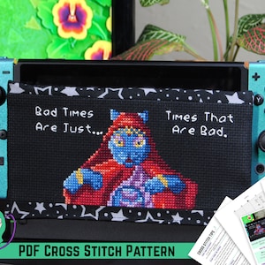 Pode incluir: Um padrão de ponto cruz para uma capa de Nintendo Switch com um personagem de gato azul com uma túnica vermelha e o texto "Bad Times Are Just... Times That Are Bad."