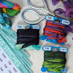 May include: Close-up of embroidery floss bobbins in various colours, including black, red, purple, green, and blue. The bobbins are labelled with the numbers 11213, 1088, and 11611. The floss is arranged on a white surface with a pair of scissors and a stack of cards labelled "Serious Stitches" with numbers 1039, 1040, 1041, 1042, 1049, 1050, 1051, 1052, 1060.