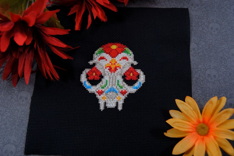 May include: A colorful embroidered skull with floral details on a black fabric background. The skull is white with red, green, blue, and yellow accents.