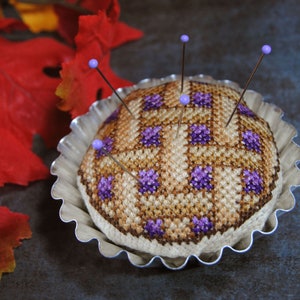 May include: A brown and purple cross-stitch pincushion in the shape of a pie, with purple beads on the pins, sitting in a silver pie pan.