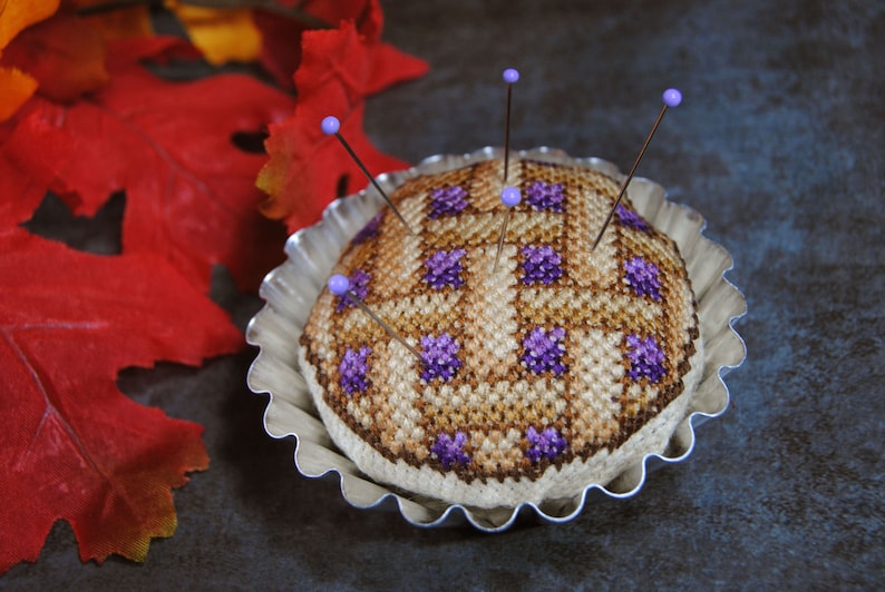 May include: A brown and purple cross-stitch pincushion in the shape of a pie, with purple beads on the pins, sitting in a silver pie pan.