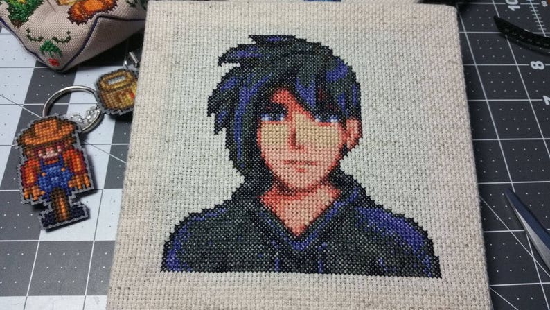 May include: Cross-stitch embroidery of a person with dark blue hair and a dark blue and green hooded shirt.