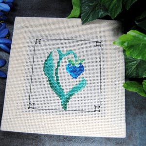 Puede incluir: Una pieza de punto de cruz cuadrada con una baya azul en forma de coraz&oacute;n y hojas verdes dentro de un cuadrado con contorno negro. La obra de arte est&aacute; sobre una tela color crema, con flores azules y hojas verdes en el fondo.
