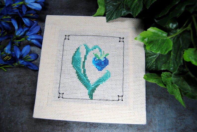 Puede incluir: Una pieza de punto de cruz cuadrada con una baya azul en forma de coraz&oacute;n y hojas verdes dentro de un cuadrado con contorno negro. La obra de arte est&aacute; sobre una tela color crema, con flores azules y hojas verdes en el fondo.
