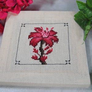 Puede incluir: Un lienzo cuadrado de color crema presenta un dise&ntilde;o de punto de cruz de una flor roja con un tallo y hojas oscuras. La flor est&aacute; enmarcada por un borde negro con detalles de esquina en forma de estrella. El lienzo est&aacute; rodeado de flores rosas y hojas verdes.