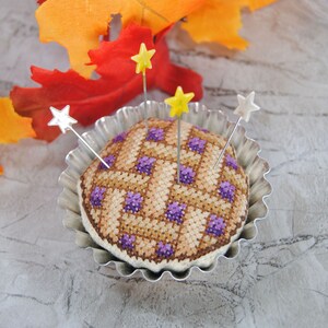 May include: A purple and brown patchwork pincushion in the shape of a pie, with four silver pins with yellow and silver star-shaped tops, in a silver pie pan.