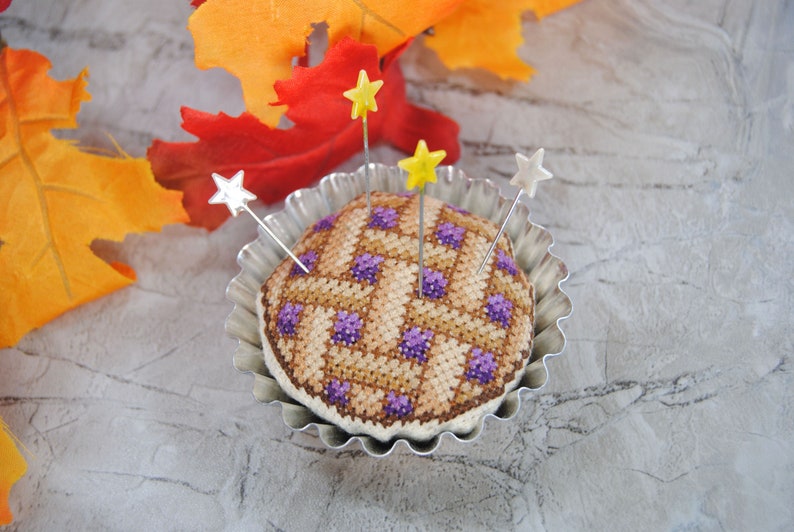 May include: A purple and brown patchwork pincushion in the shape of a pie, with four silver pins with yellow and silver star-shaped tops, in a silver pie pan.