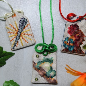 May include: Three embroidered Japanese omamori charms with different designs. One features a sunburst, another a bird on a branch, and the third a figure holding an object. Each charm is attached to a cord.