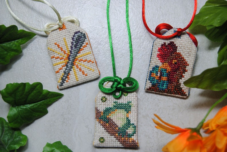 May include: Three embroidered Japanese omamori charms with different designs. One features a sunburst, another a bird on a branch, and the third a figure holding an object. Each charm is attached to a cord.