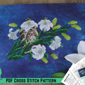 Pode incluir: Um padrão de ponto cruz com um pássaro castanho aninhado em um buquê de lírios brancos com folhas verdes sobre um fundo azul. O padrão é intitulado "Grandma's Woods" e inclui um padrão de ponto cruz PDF.