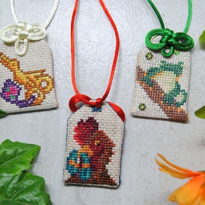 May include: Three embroidered Japanese omamori charms with different designs. One features a gold and purple design, another a red and orange figure, and the third a green and brown design. Each charm is attached to a cord.