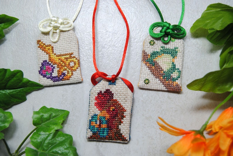 May include: Three embroidered Japanese omamori charms with different designs. One features a gold and purple design, another a red and orange figure, and the third a green and brown design. Each charm is attached to a cord.