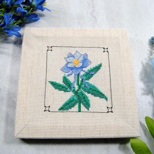 Puede incluir: Una pieza cuadrada de punto de cruz blanquecina con una flor azul con centro amarillo y hojas verdes, enmarcada en negro. La pieza est&aacute; rodeada de flores azules.