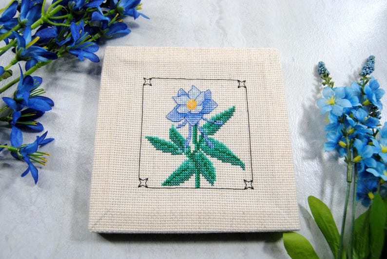 Puede incluir: Una pieza cuadrada de punto de cruz blanquecina con una flor azul con centro amarillo y hojas verdes, enmarcada en negro. La pieza est&aacute; rodeada de flores azules.