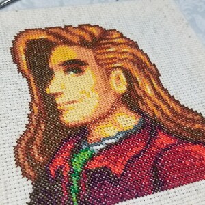 May include: Cross-stitch embroidery of a person with long brown hair, wearing a red and green jacket. The person is looking to the right.