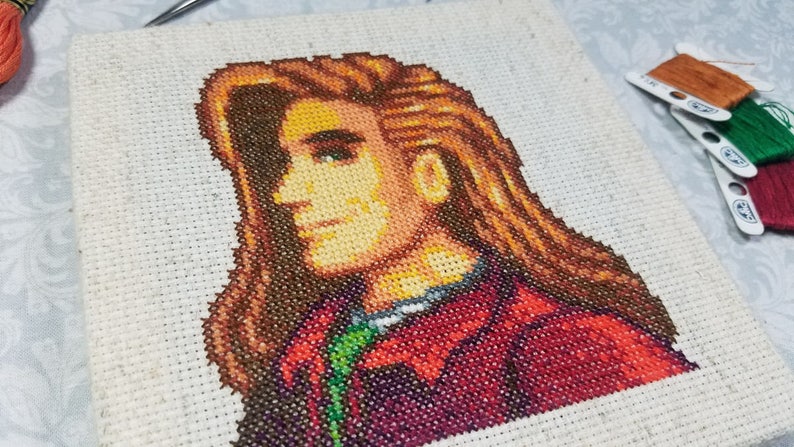 May include: Cross-stitch embroidery of a person with long brown hair, wearing a red and green jacket. The person is looking to the right.