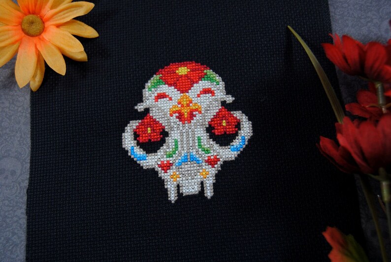 May include: A colorful cross-stitch design of a sugar skull with red, white, blue, green, and yellow embroidery floss. The skull is smiling and has flowers on the top of its head.