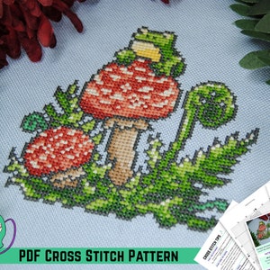 Può includere: Un modello a punto croce con un design stravagante di un fungo con cappello rosso con punti bianchi, un fungo più piccolo e una rana verde. Il design include fogliame verde e il testo "PDF CROSS STITCH PATTERN".