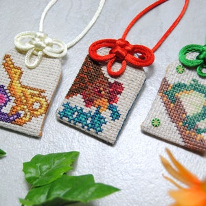 May include: Three embroidered Japanese omamori charms with colorful designs. The charms are rectangular with a cream-colored background and feature intricate cross-stitch patterns. Each charm has a different colored cord and knot.