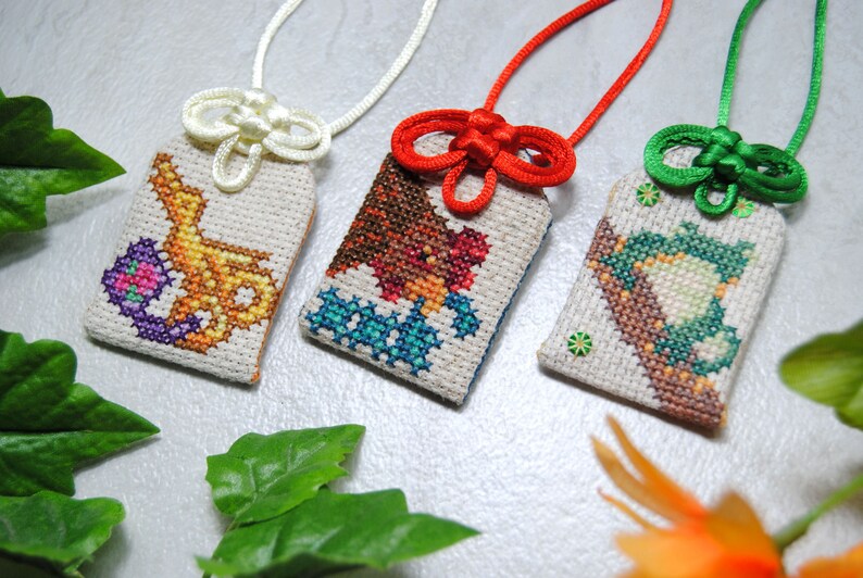 May include: Three embroidered Japanese omamori charms with colorful designs. The charms are rectangular with a cream-colored background and feature intricate cross-stitch patterns. Each charm has a different colored cord and knot.