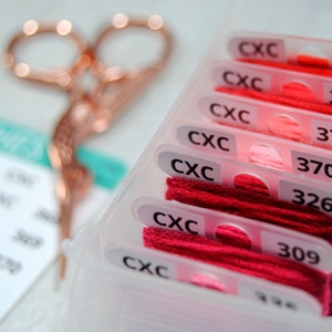 May include: A clear plastic container with rows of colorful embroidery floss. Each floss is labeled with a number and the letters "CXC". The floss colors range from pink to red.