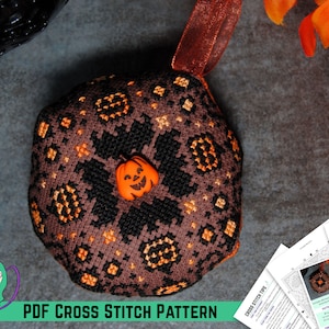 以下が含まれることがあります： 茶色と黒のクロスステッチパターンで、ジャック・オ・ランタンの顔をしたカボチャが描かれています。パターンは、茶色のリボンが付いた丸いクッションに施されています。クッションは灰色の表面に置かれています。画像には、印刷されたPDFクロスステッチパターンと説明書も含まれています。