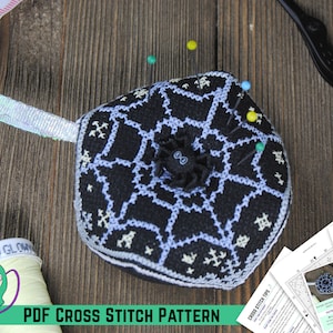 May include: A black and white cross stitch pattern of a spider web with a spider in the center. The pattern is on a hexagonal pincushion with a white ribbon loop. The pincushion is surrounded by sewing supplies, including scissors, pins, and a spool of thread.