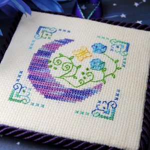 May include: Square cross-stitch embroidery featuring a crescent moon in purple and blue hues, with a yellow butterfly and blue flowers. The design is framed by a dark purple cord and corner accents. The background is a starry night.
