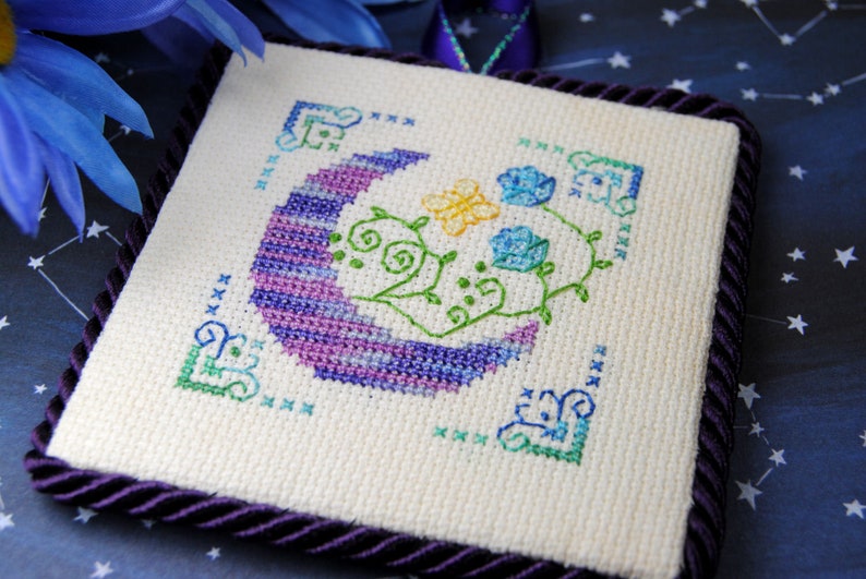 May include: Square cross-stitch embroidery featuring a crescent moon in purple and blue hues, with a yellow butterfly and blue flowers. The design is framed by a dark purple cord and corner accents. The background is a starry night.