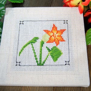 Puede incluir: Un cuadrado blanco de punto de cruz con una flor naranja y hojas verdes, enmarcado por un borde negro. El bordado representa un dise&ntilde;o floral. Es probable que sea un art&iacute;culo artesanal hecho a mano.