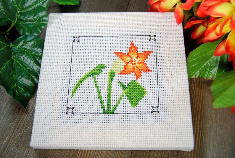Puede incluir: Un cuadrado blanco de punto de cruz con una flor naranja y hojas verdes, enmarcado por un borde negro. El bordado representa un dise&ntilde;o floral. Es probable que sea un art&iacute;culo artesanal hecho a mano.