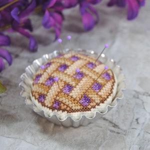 May include: A purple and brown cross-stitch pincushion in the shape of a pie, with purple pins, sitting in a silver pie pan.